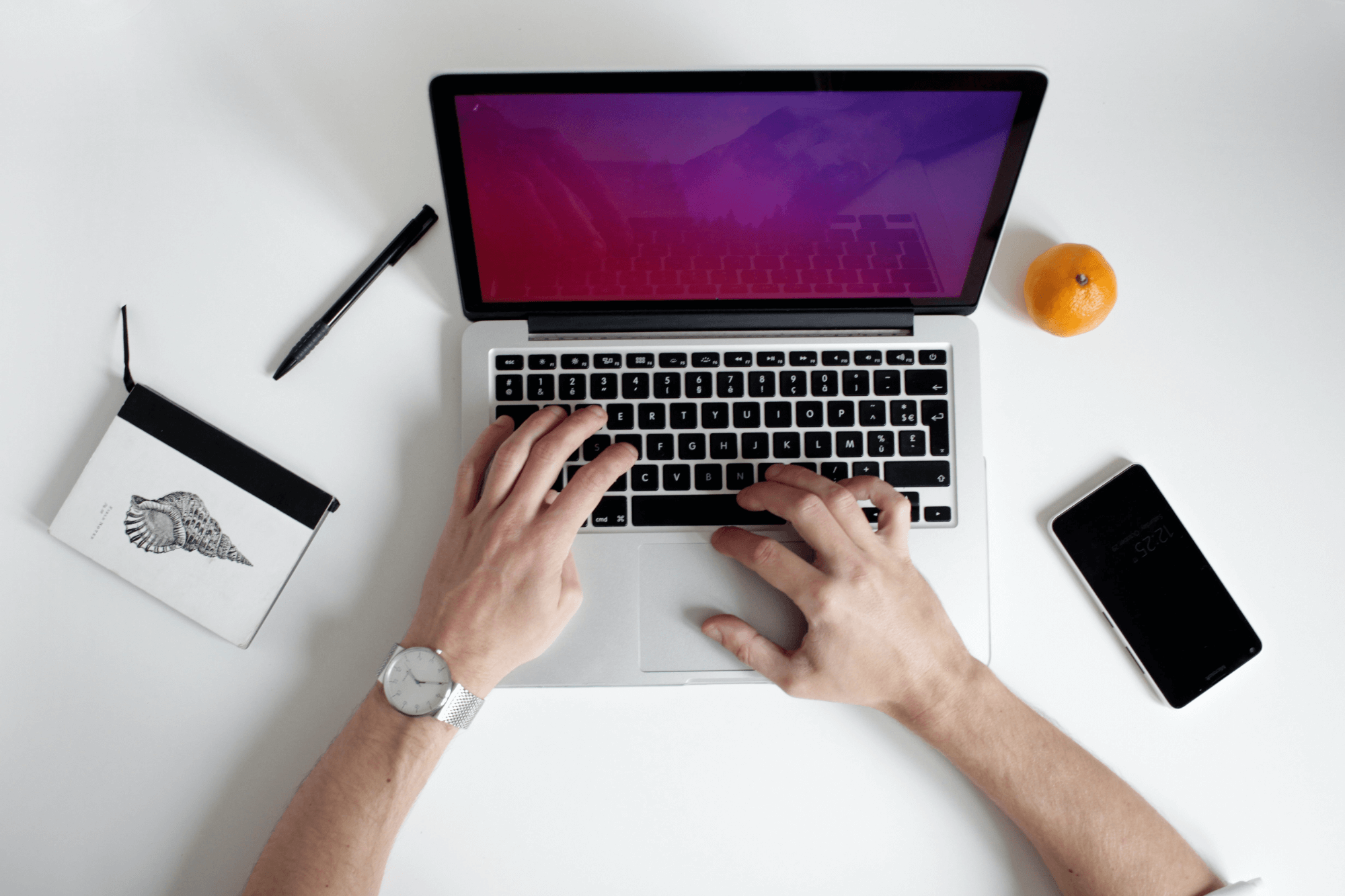 visão de cima de uma área de trabalho. No centro, há um laptop com uma tela de descanso roxa e as mãos de alguém digitando nele. À esquerda do laptop, há um caderno branco com uma ilustração de uma cabeça de zebra na capa e uma caneta preta diagonalmente sobre ele. À direita, há uma laranja e, mais abaixo, um smartphone. A pessoa que está trabalhando usa um relógio prateado no pulso esquerdo.