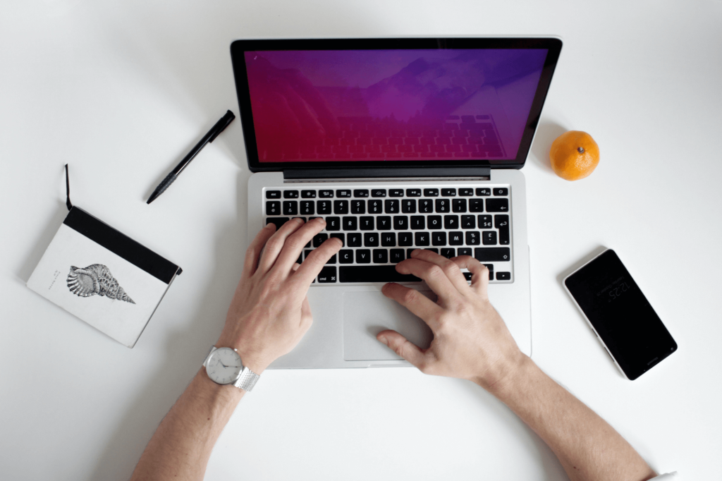 Visão de cima de uma área de trabalho com um laptop no centro, mãos digitando, um caderno e uma caneta à esquerda, uma laranja e um smartphone à direita. A pessoa usa um relógio no pulso esquerdo