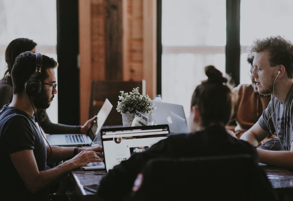 A imagem mostra um grupo de pessoas sentadas em uma mesa, trabalhando em laptops com rostos pixelados para preservar a privacidade. Uma planta em vaso adiciona um toque de verde ao ambiente, que parece um espaço de co-working moderno e colaborativo.