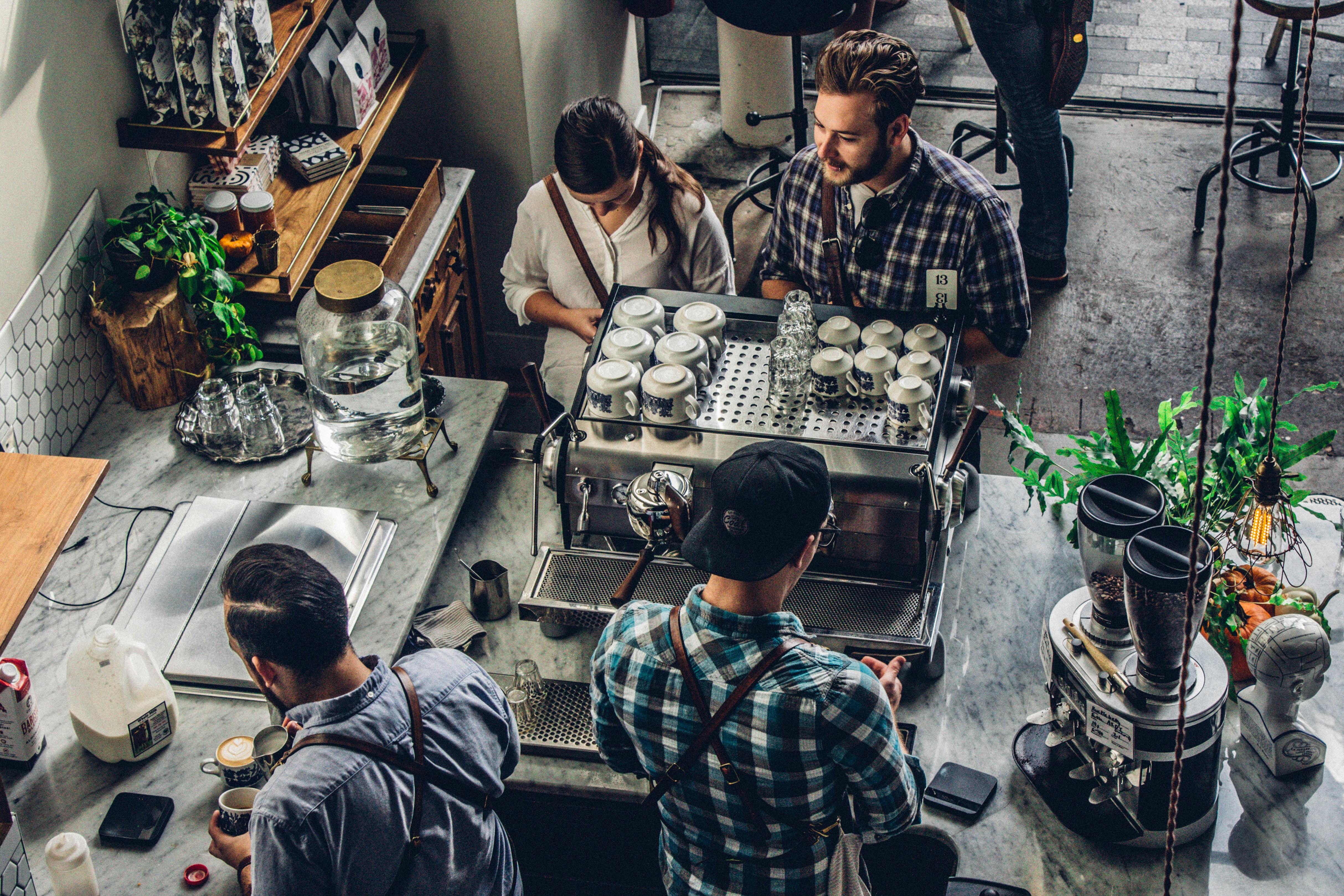 Visão aérea de uma cena em uma cafeteria. Quatro pessoas estão ao redor de um balcão de café equipado com máquinas. Uma pessoa parece estar preparando uma bebida.